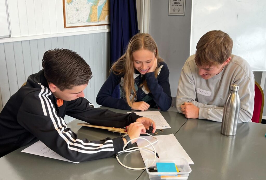 Image of three students working collaboratively at a table, paper and cables on table. They are smiliing.